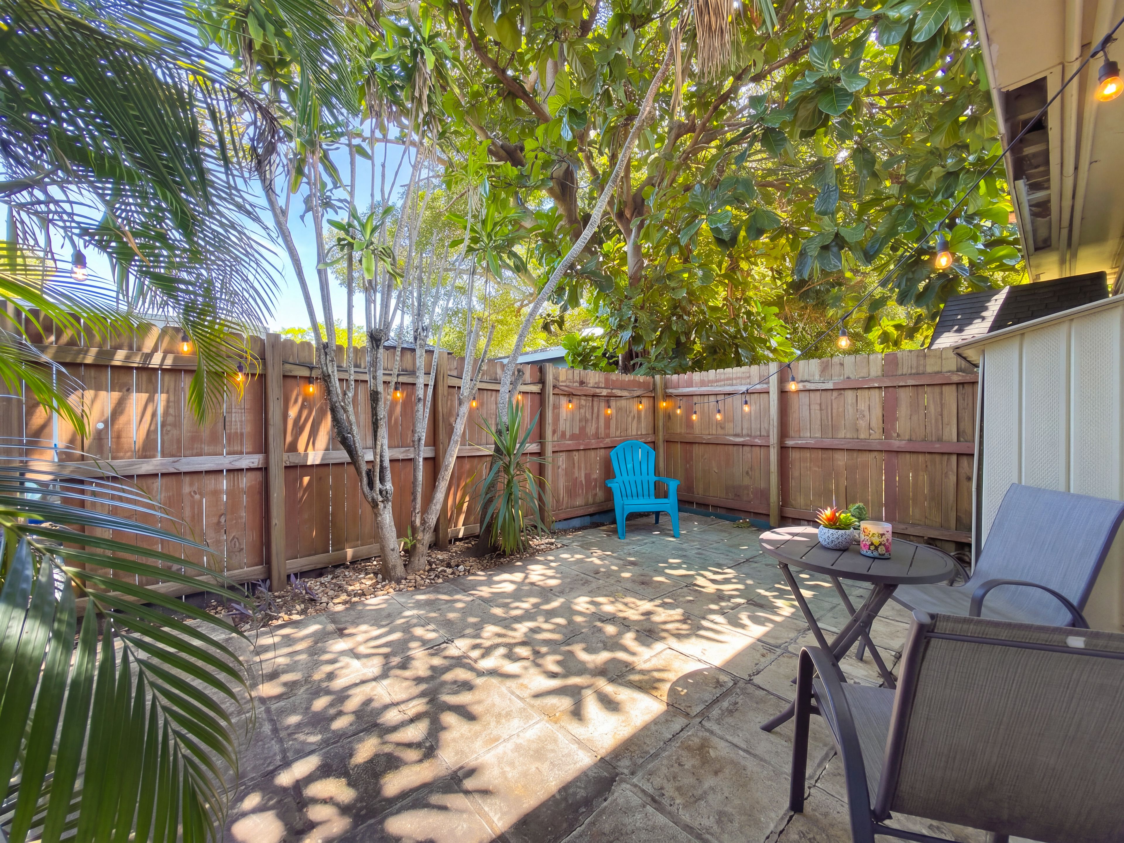 A backyard with a wooden fence, a blue chair, a table with a candle, and a potted plant.