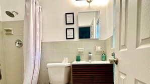 A bathroom with a toilet, sink, and shower. There are framed pictures on the wall and a mirror above the sink.