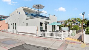 A modern building with a white exterior, a balcony with a striped umbrella, and a gated patio area. - Mission Paradise - Spa, Pet Friendly, Steps to Sand & W/D! (Mission Beach)