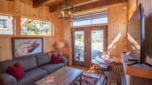 Ein gemütliches Blockhaus-Interieur mit Holzausstattung, einer Couch mit roten Kissen, einem Couchtisch, einem Esstisch mit Stühlen und einem an der Wand befestigten Fernseher.
