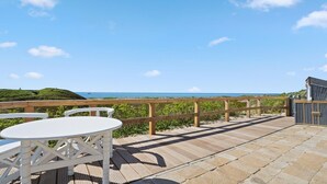 Une terrasse en bois avec une table et des chaises blanches, donnant sur une plage et l’océan.