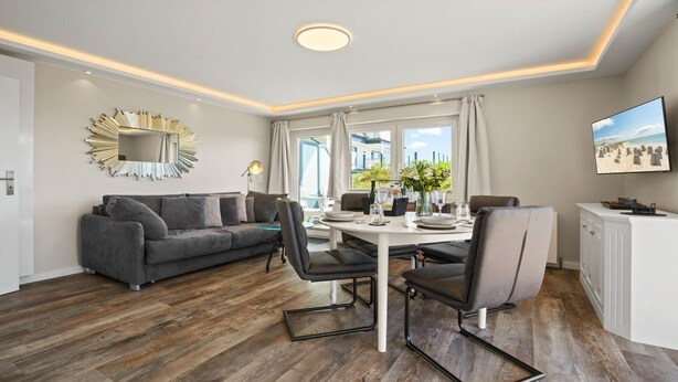 Un salon moderne avec un canapé gris, une table à manger avec des chaises, un téléviseur fixé au mur et une grande fenêtre avec des rideaux.