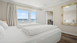 A hotel room with a large bed, a window with curtains, a mirror, and a view of the city. - Käpitäns-suite - Haus Seeblick (Hörnum (Sylt))