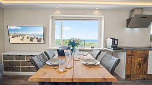 A dining area with a wooden table set for four, a view of the beach, and a kitchen area with a modern stove. - Käpitäns-suite - Haus Seeblick (Hörnum (Sylt))