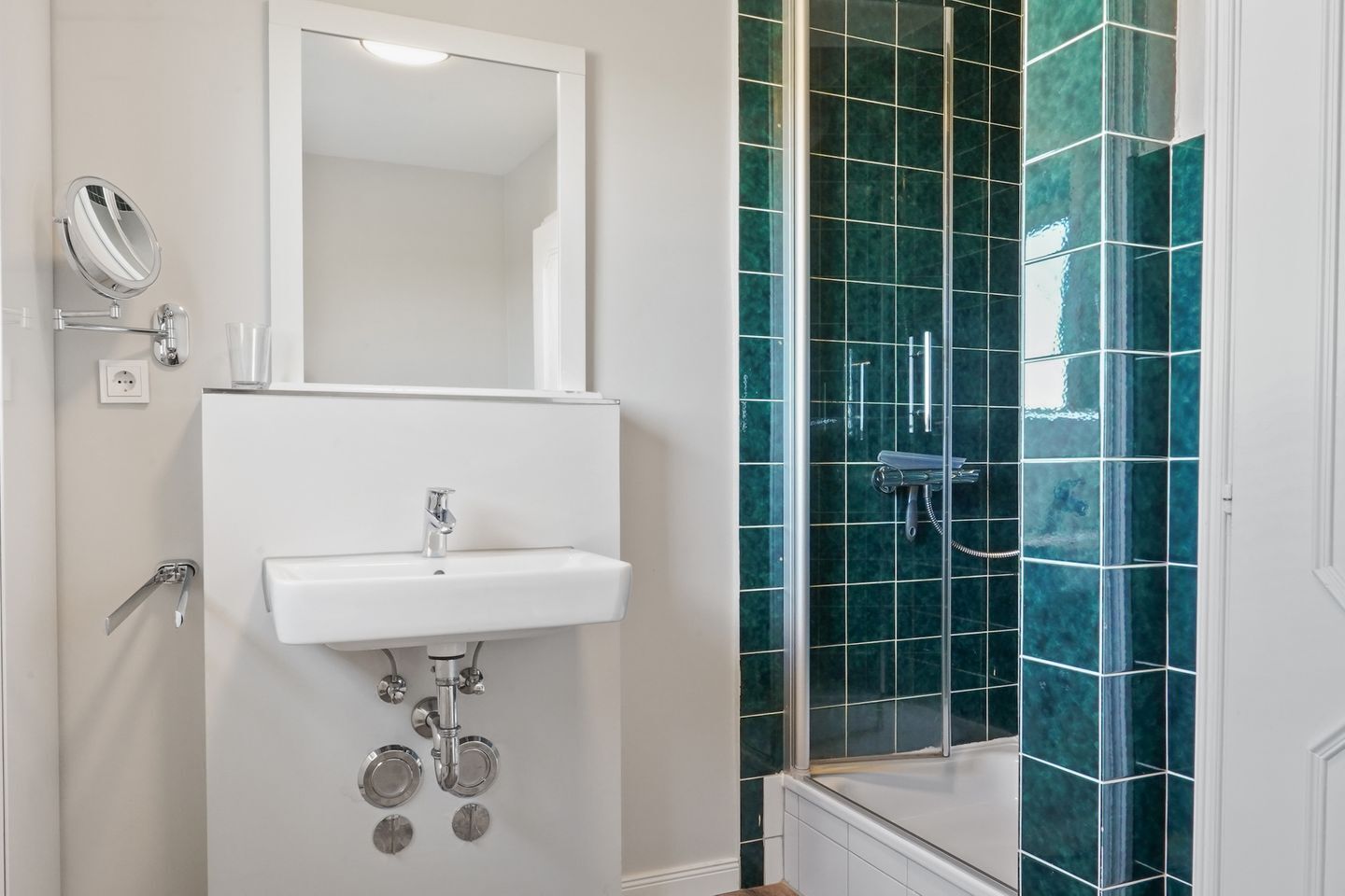 A bathroom with a white sink, a mirror, and a shower with green tiles.