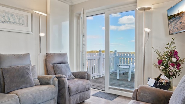 A room with two grey armchairs, a wooden floor, a white ceiling, a large window with a view of a white fence and sky, and a wall-mounted TV. - Meerluft-suite - Haus Seeblick (Hörnum (Sylt))
