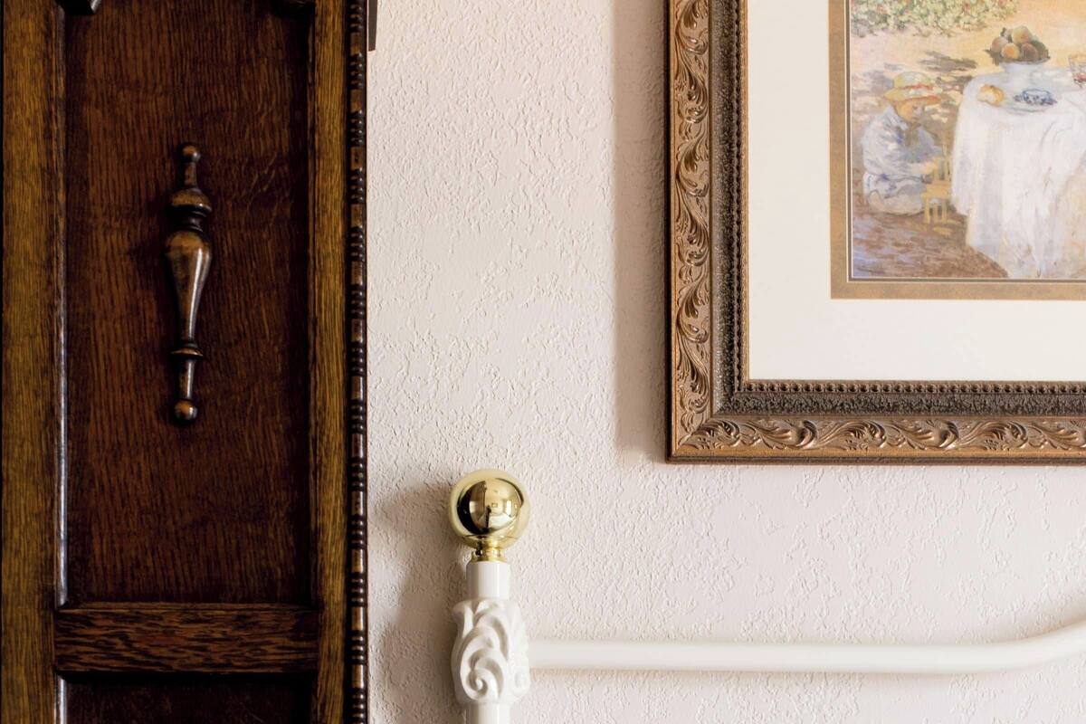 A white bed with a gold knob, a wooden wardrobe, and a framed painting on the wall.