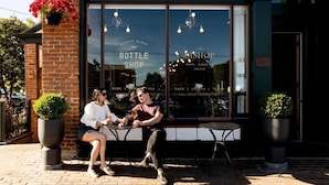 A brick building with a large window displaying the text "BOTTLE SHOP" and "BISHOP BLOCK HOTEL CHECK-IN". There are two people sitting at an outdoor table, and potted plants are visible on the sidewalk. - The Bishop Hotel (Port Townsend)