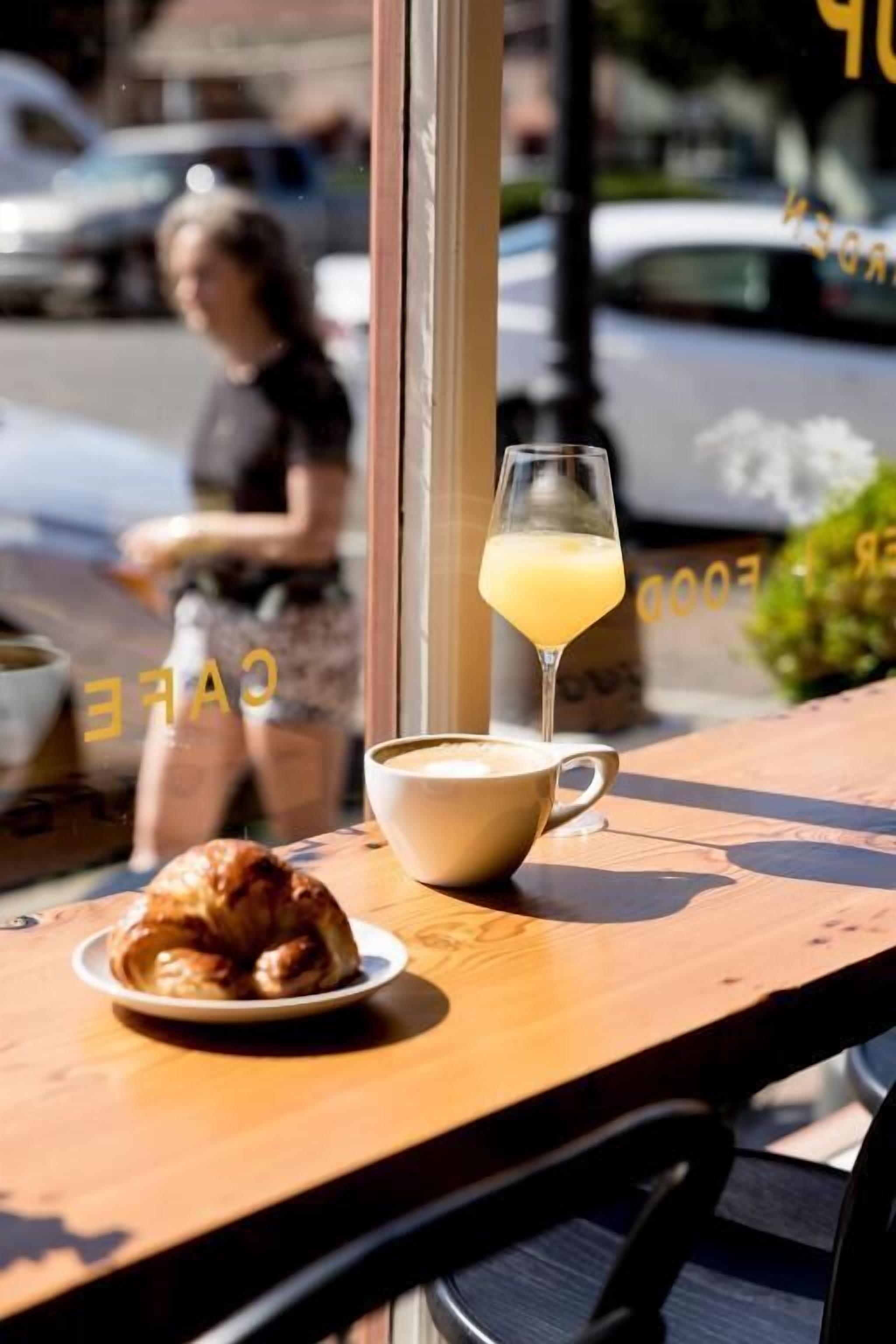 A table with a croissant and a cup of coffee, a glass of orange juice, and a view of the street outside.