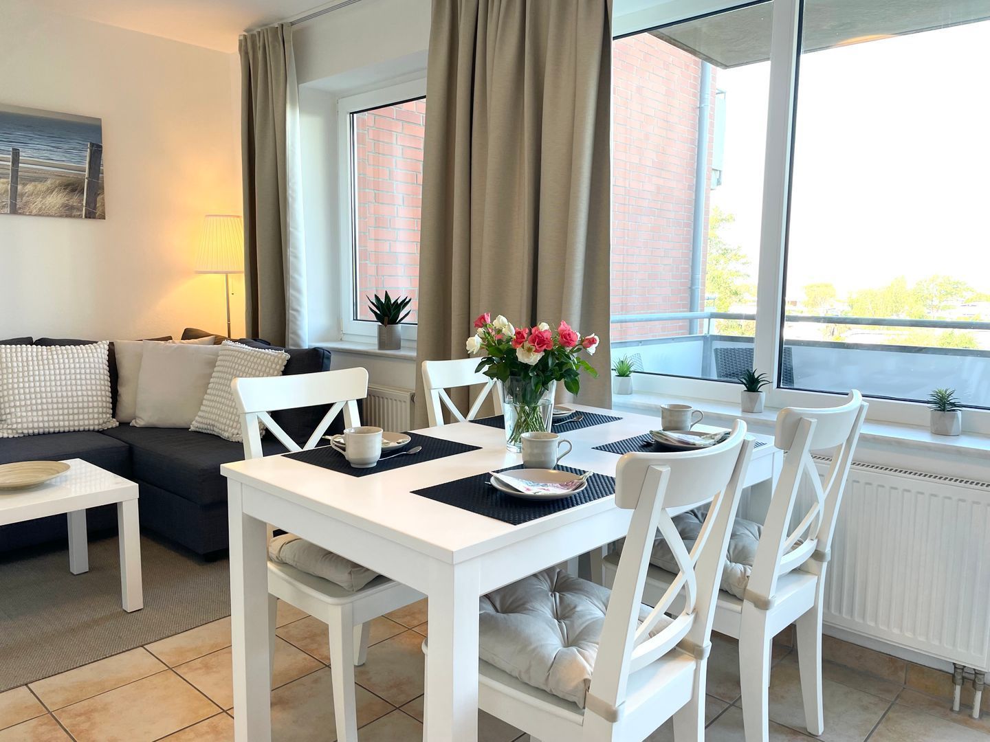 A modern living room with a dining area, featuring a white table and chairs, a sofa, and a large window with curtains.
