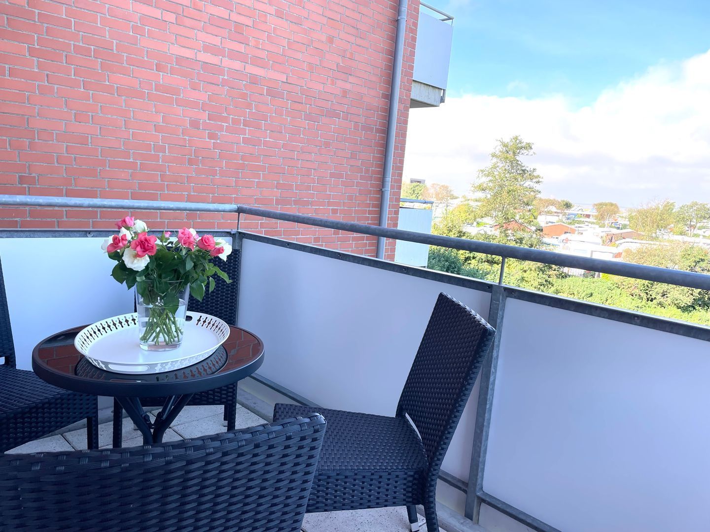Ein Balkon mit Tisch und Stühlen, einem Blumenstrauß und Blick auf ein Backsteinhaus sowie Grünflächen.