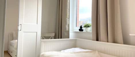A bedroom with a bed, a window with curtains, a radiator, and a small potted plant on the windowsill.