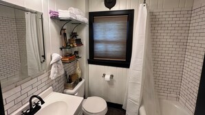 Un baño pequeño con un lavabo, inodoro y zona de ducha de color blanco. Hay una estantería con libros y objetos decorativos, y una ventana con persianas.