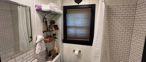 A compact bathroom with a white sink, toilet, and shower area. There is a shelf with books and decorative items, and a window with blinds.