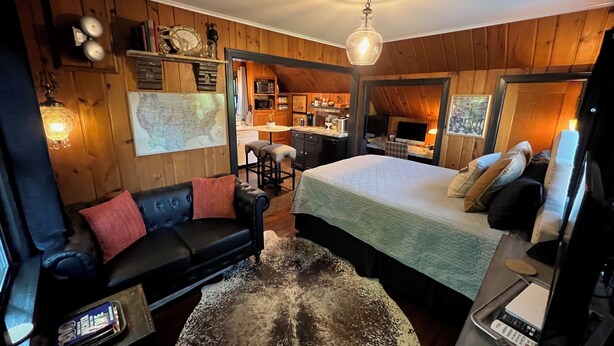 Una habitación acogedora con una cama, un sofá de cuero negro, techo de madera y una cocina visible a través de una puerta abierta.