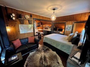 Una habitación acogedora con una cama, un sofá de cuero negro, techo de madera y una cocina visible a través de una puerta abierta.