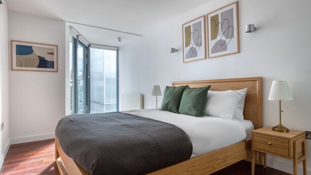 Un dormitorio moderno con cama de madera, almohadas verdes y blancas, una mesita de noche con lámpara y un ventanal con vistas.