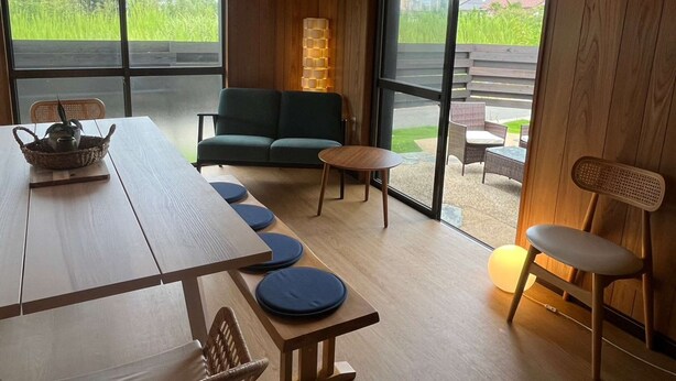 Un interior moderno con una mesa de madera, asientos acolchados azules, un sofá verde y ventanas amplias que ofrecen vistas a la vegetación y un edificio.