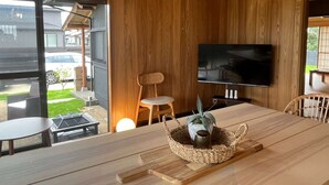 Una habitación moderna con paredes de madera, un televisor grande montado en la pared, una mesa de comedor con sillas y un pequeño jardín visible a través de la puerta de vidrio.