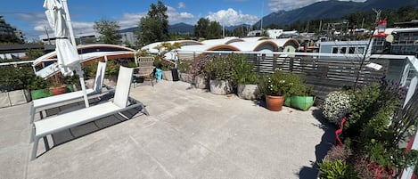 A rooftop terrace with potted plants, a white chair, and a view of boats and mountains.