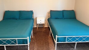 A room with two single beds, each with a teal mattress and matching pillows, a white bedside table with drawers, and a carpeted floor.