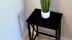 A small black table with a white pot containing green plants, positioned against a plain wall with a light switch above it.