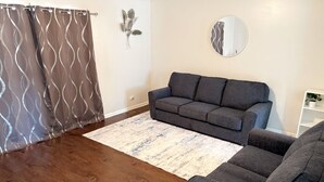 A living room with a grey sofa, a patterned rug, wooden flooring, a grey curtain, and a wall-mounted mirror.