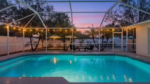Una piscina con un porche cubierto, rodeada por una cerca blanca y luces colgantes.