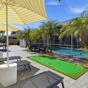 A poolside area with a mini-golf setup, outdoor seating, and a large striped umbrella. - Free Heated Pool, Baby Gear, Shuffleboard, EV charger, Near beach, UTC mall, SRQ (Sarasota)