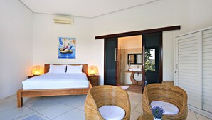 A bedroom with a large bed, two wicker chairs, a small table with a vase, and a painting on the wall. - Villa Kipas Retreat (Senggigi)