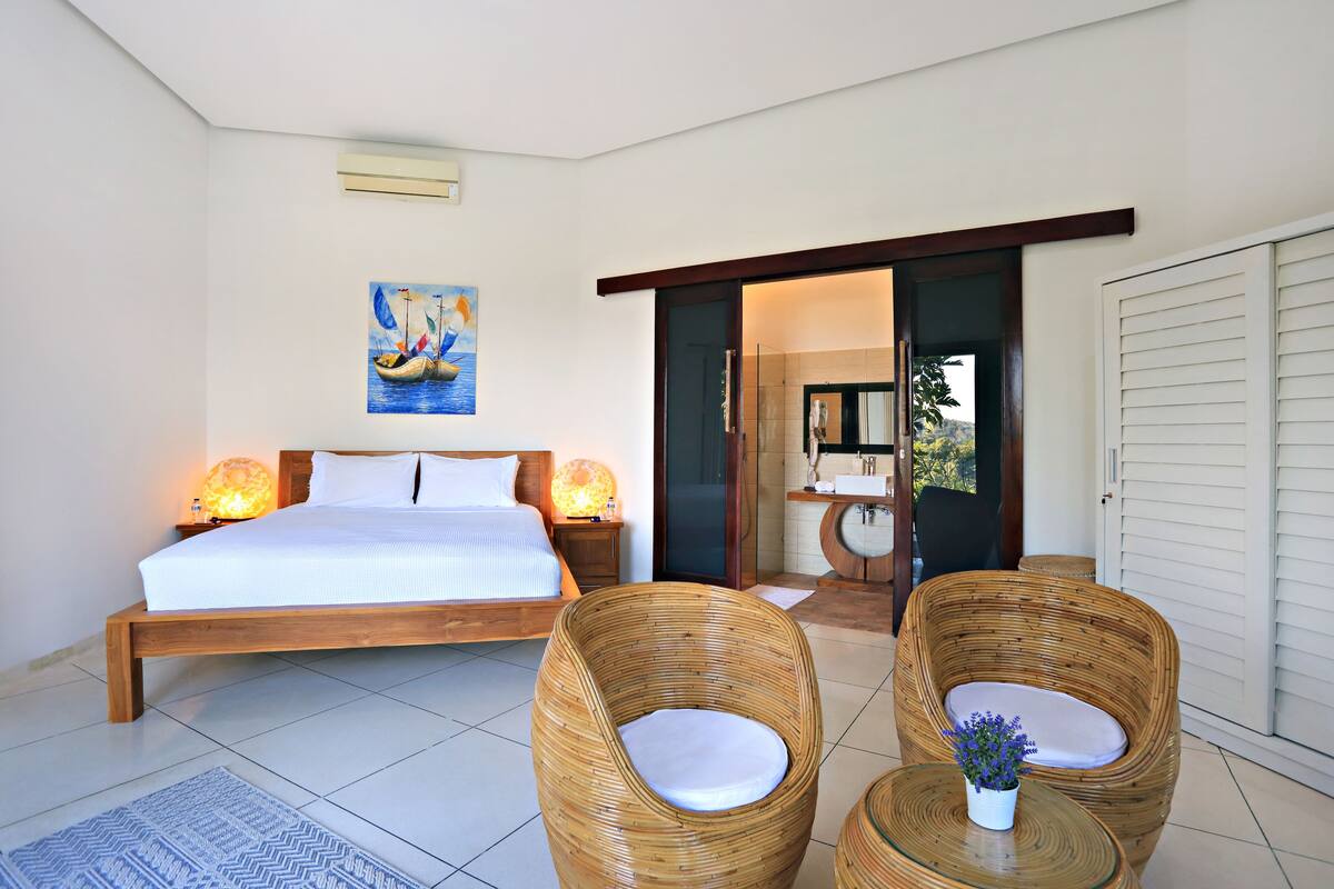 A bedroom with a large bed, two wicker chairs, a small table with a vase, and a painting on the wall.