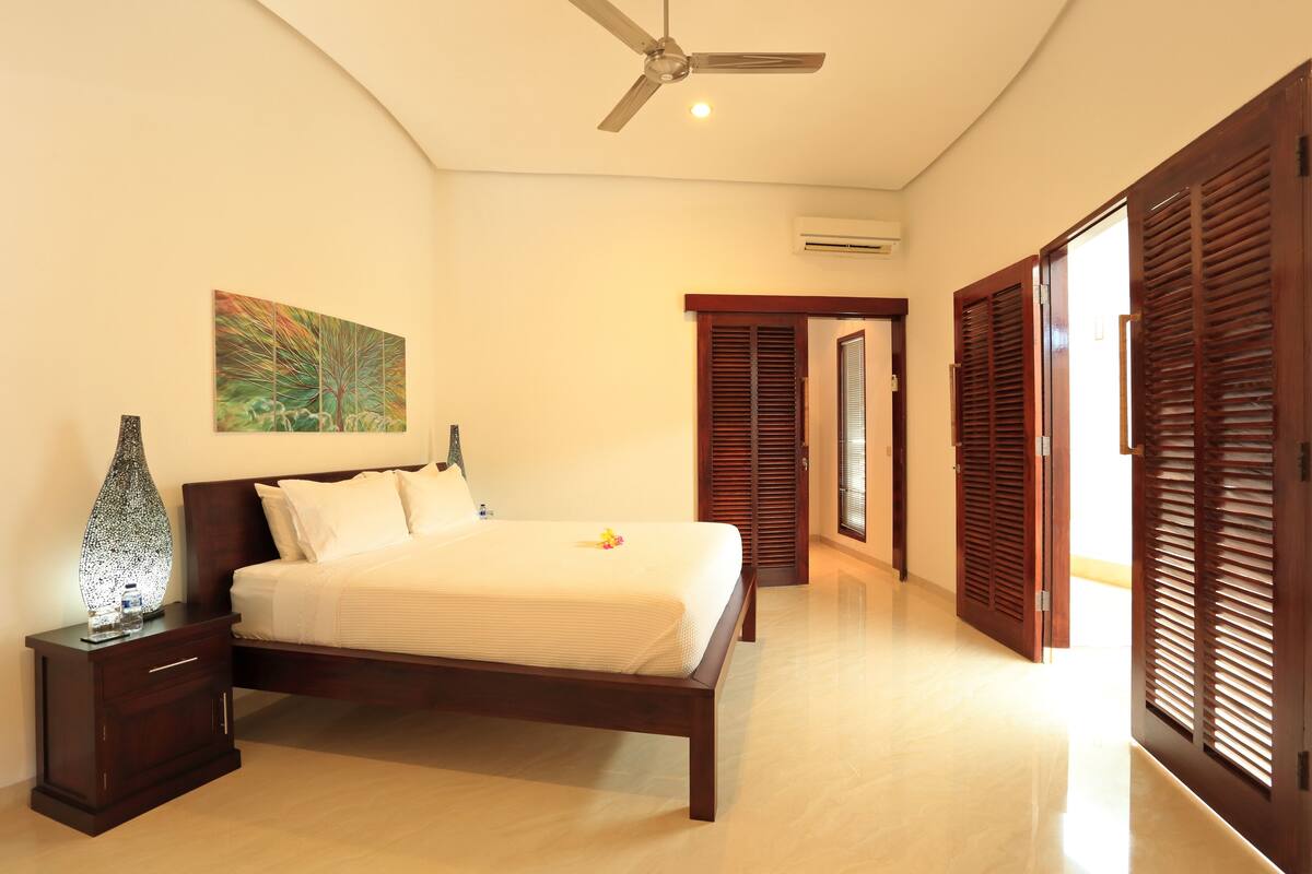 A bedroom with a bed, nightstand, ceiling fan, artwork, and wooden shutters.