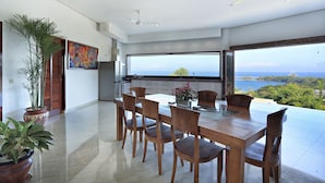 A modern dining area with a large table and chairs, a ceiling fan, and a view of the ocean. - Villa Kipas Retreat (Senggigi)