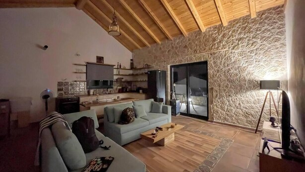 A spacious living area with a stone wall, wooden ceiling, and a kitchen in the background. - Villa with pool in Pelekas near the beach (Pelekas)