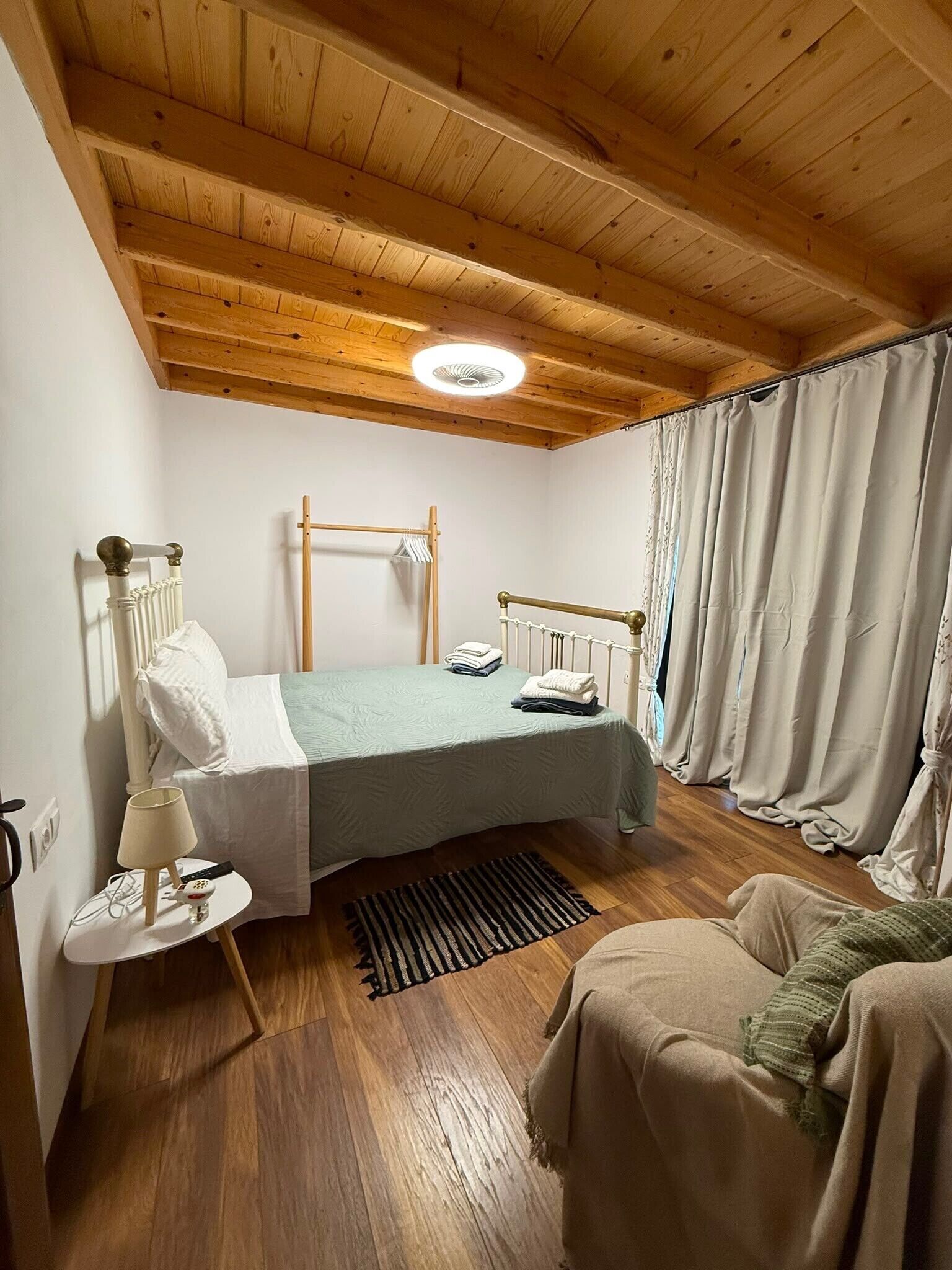 A bedroom with a wooden ceiling, a bed with a headboard, a bedside table with a lamp, and a window with curtains.