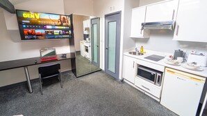 Une chambre d’hôtel moderne équipée d’un téléviseur à écran plat, d’un bureau avec un ordinateur portable, d’une petite chaise, d’une kitchenette avec un four à micro-ondes, un four et un évier, et d’une porte vitrée donnant accès à la salle de bain.