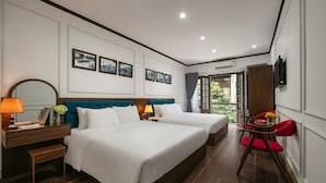 Une chambre d’hôtel avec deux lits, un bureau avec une lampe, un miroir et une vue sur la verdure par une porte ouverte.