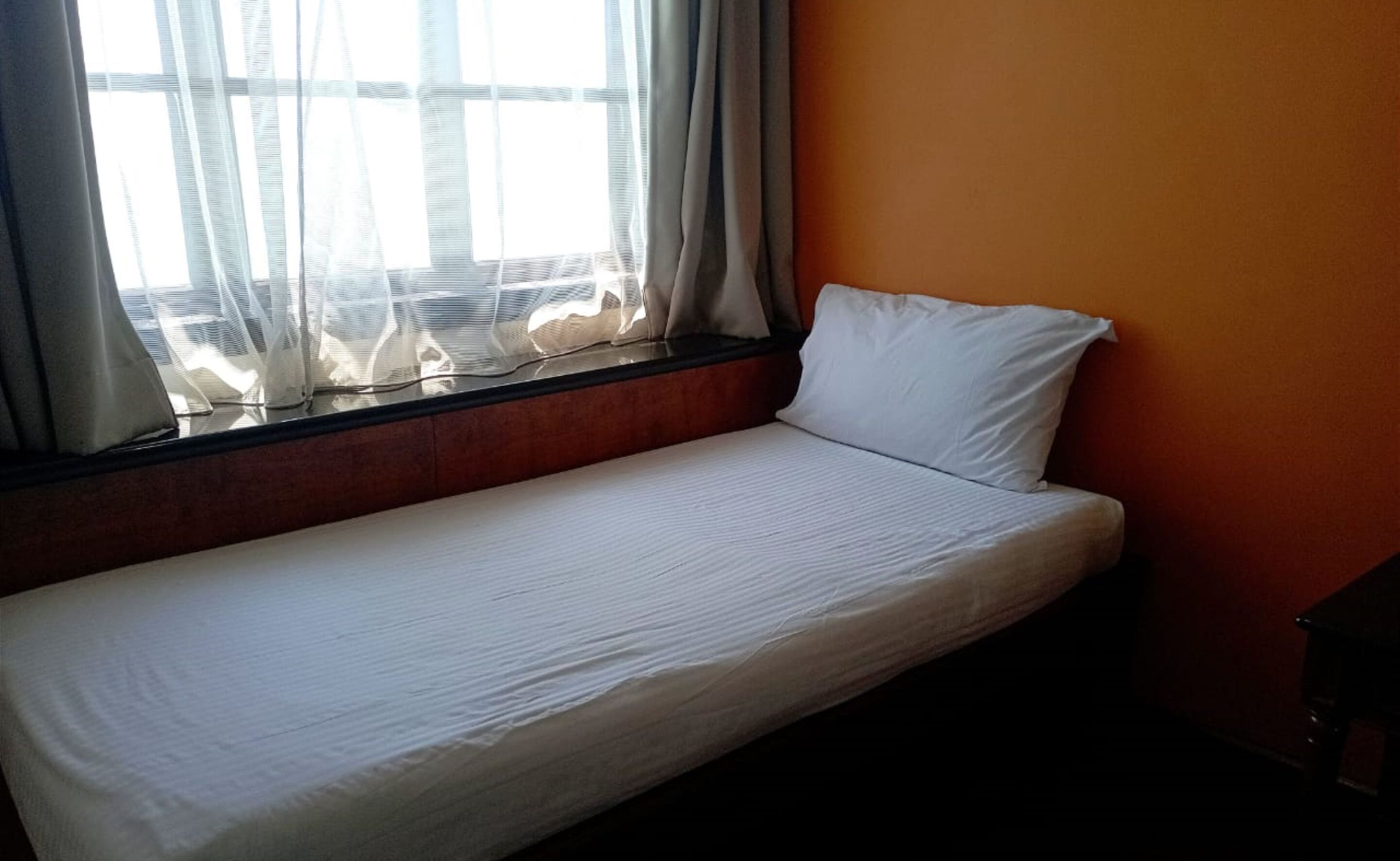 Una cama individual en una habitación con ventana, cortina y pared naranja.