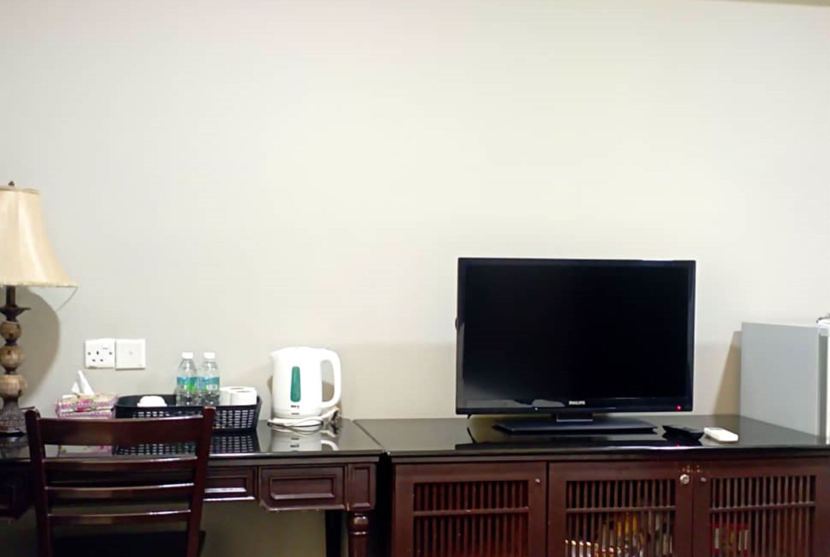 A hotel room with a desk, chair, television, lamp, and a cabinet with bottles.