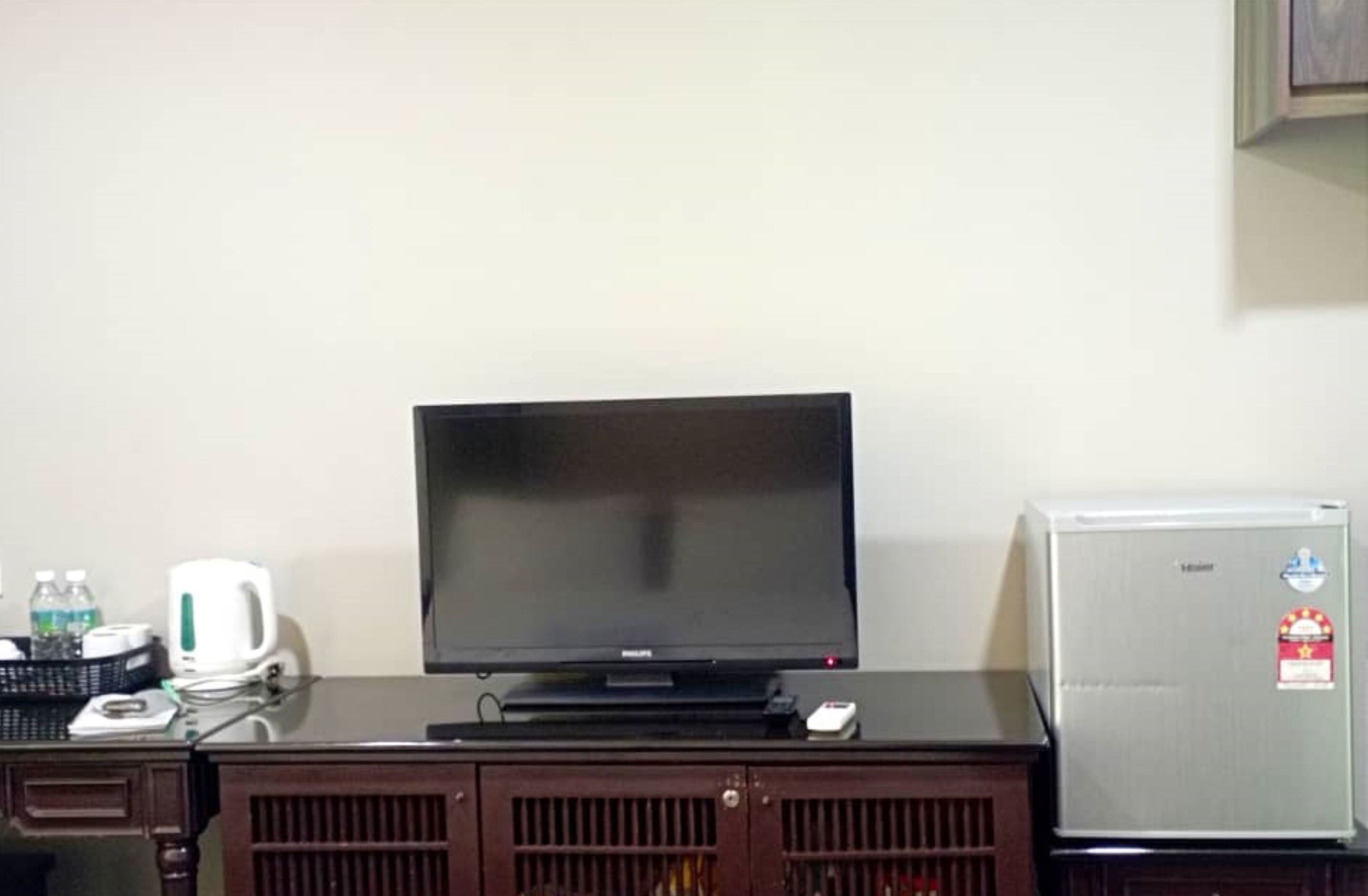 A television on a wooden cabinet, a refrigerator, and a wall-mounted shelf with a kettle and bottles.