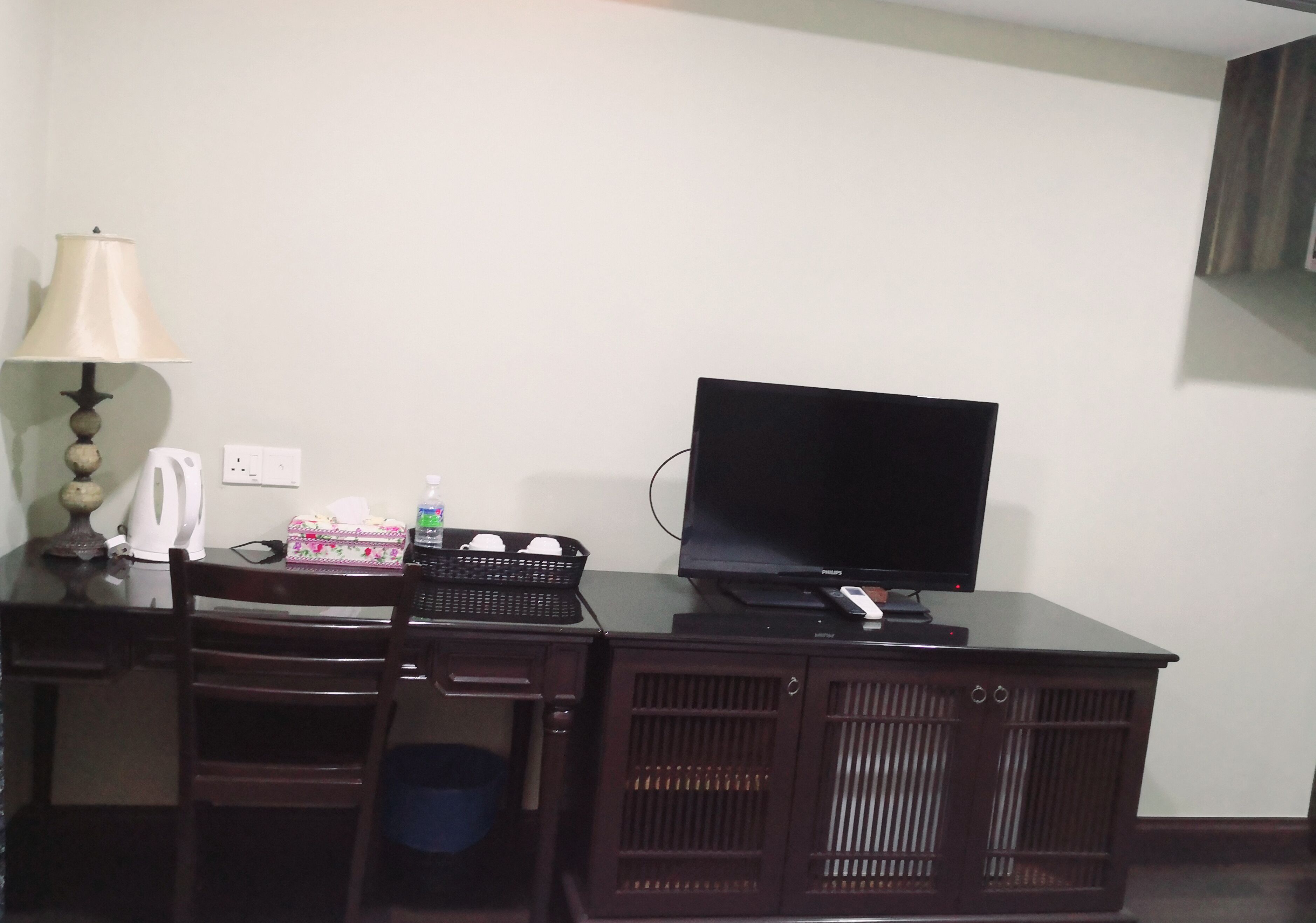 A hotel room with a wooden desk, a flat-screen TV, a lamp, a kettle, a box of tissues, and a bottle of water.