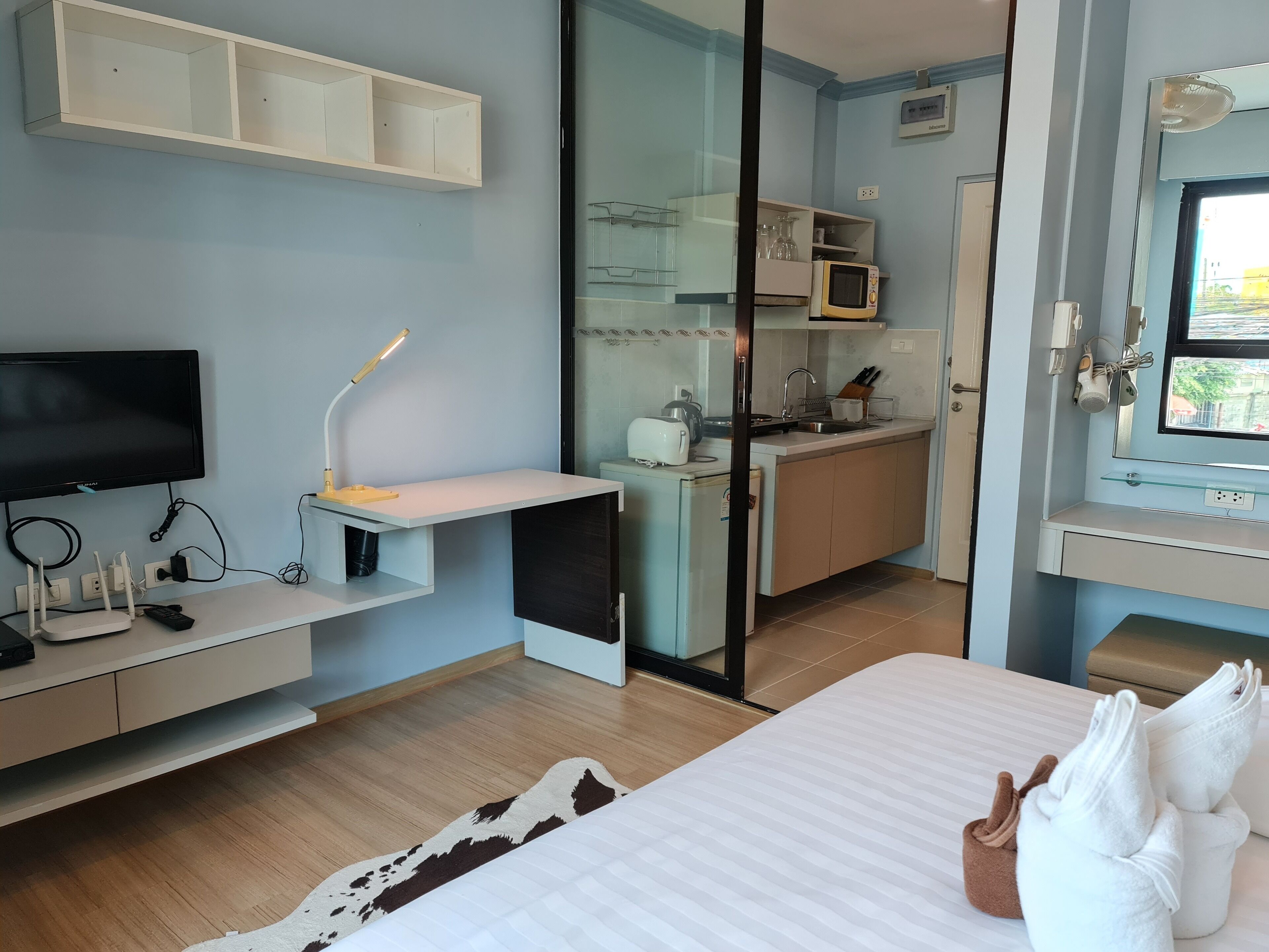A modern hotel room with a flat-screen TV, desk with lamp, kitchenette, and a bed with towels.