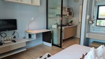 A modern hotel room with a flat-screen TV, desk with lamp, kitchenette, and a bed with towels.