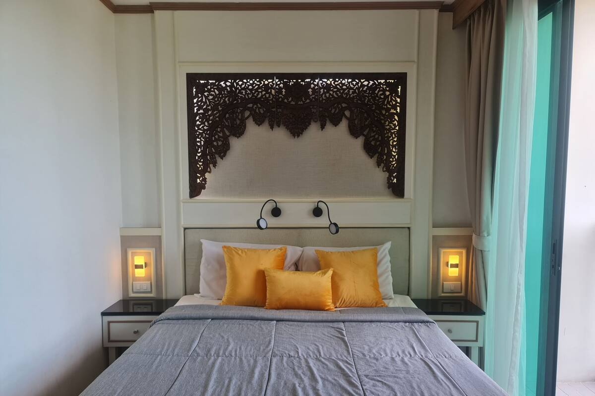 Una cama bien hecha con edredón gris y dos almohadas amarillas, flanqueada por mesitas de noche con lámparas.