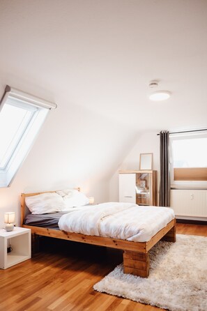 A bedroom with a sloped ceiling, a wooden bed, a nightstand, a white rug, and a window with curtains.