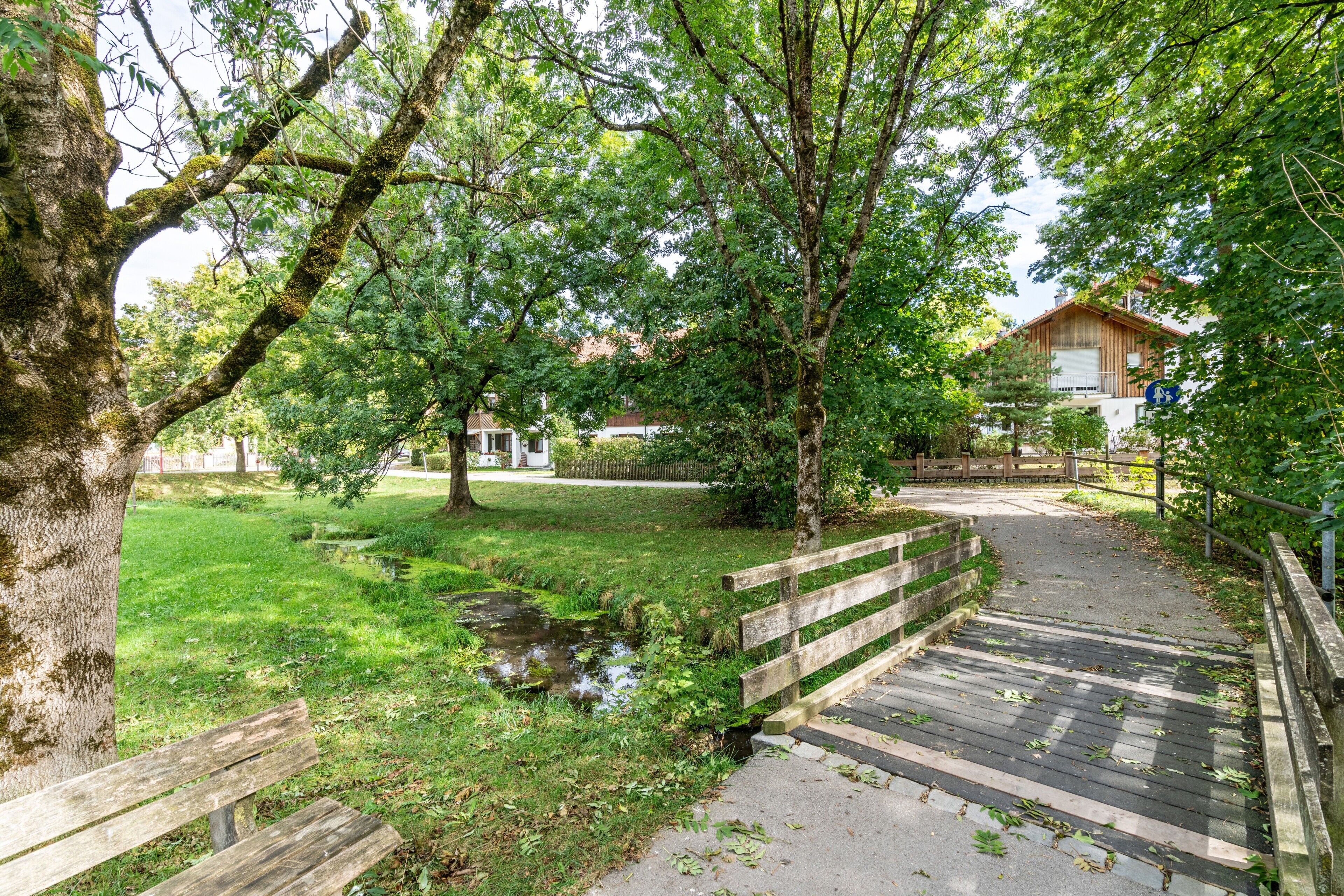 Un sentier dans un parc, avec un banc en bois, un petit ruisseau et un bâtiment en arrière-plan.