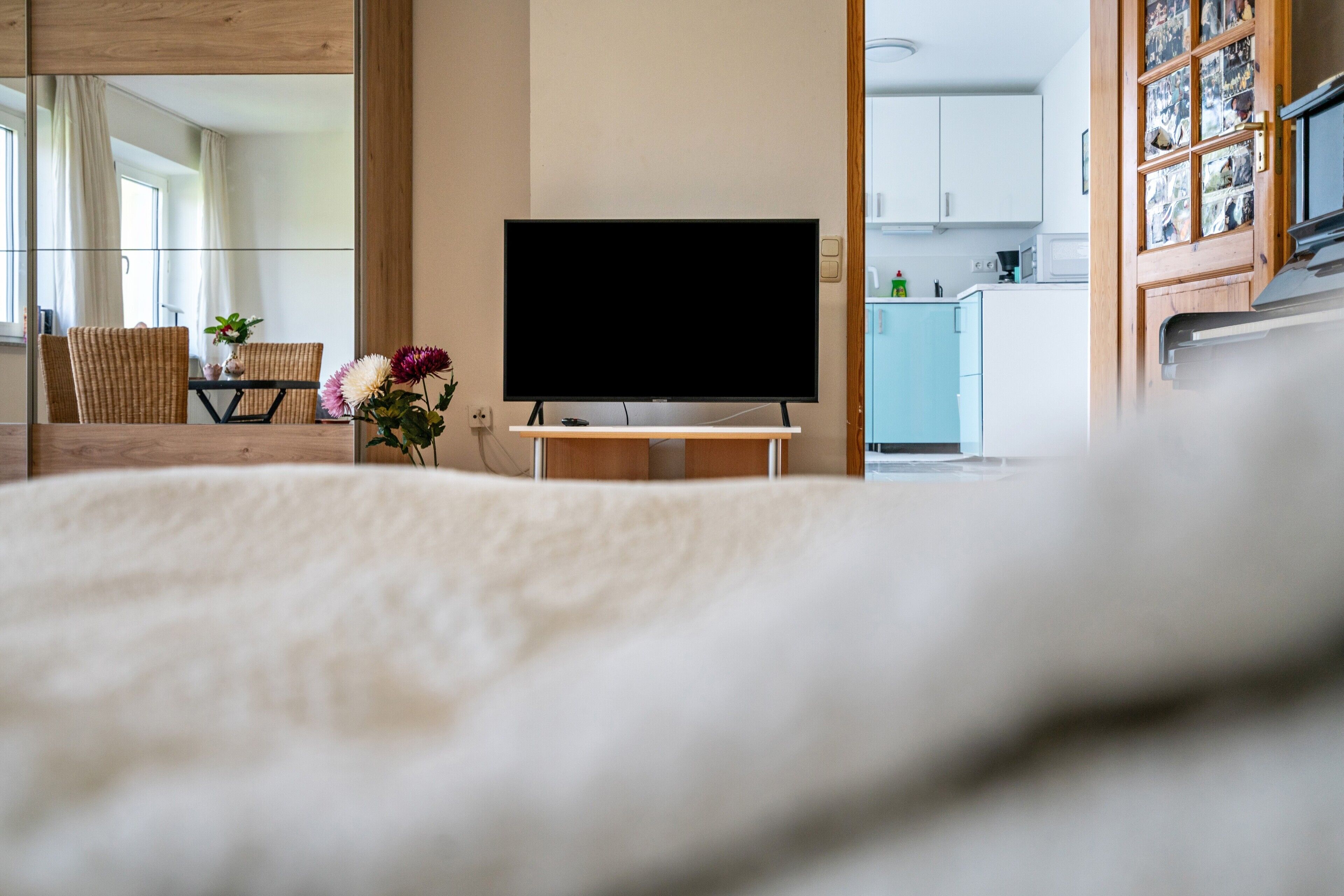 Une chambre avec un lit, un téléviseur posé sur un meuble, un coin repas avec des chaises et une table, et une cuisine visible à travers une porte ouverte.