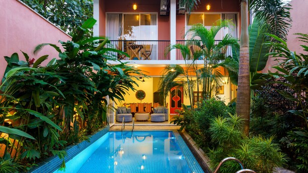 Una piscina con borde de azulejos, rodeada de exuberante vegetación y un edificio con balcón.