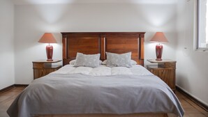 Una cama bien hecha con cabecera de madera, dos mesitas de noche con lámparas y una ventana a la derecha.