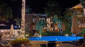 Zona de piscina nocturna con sillones de descanso, piscina y un edificio con varios balcones.